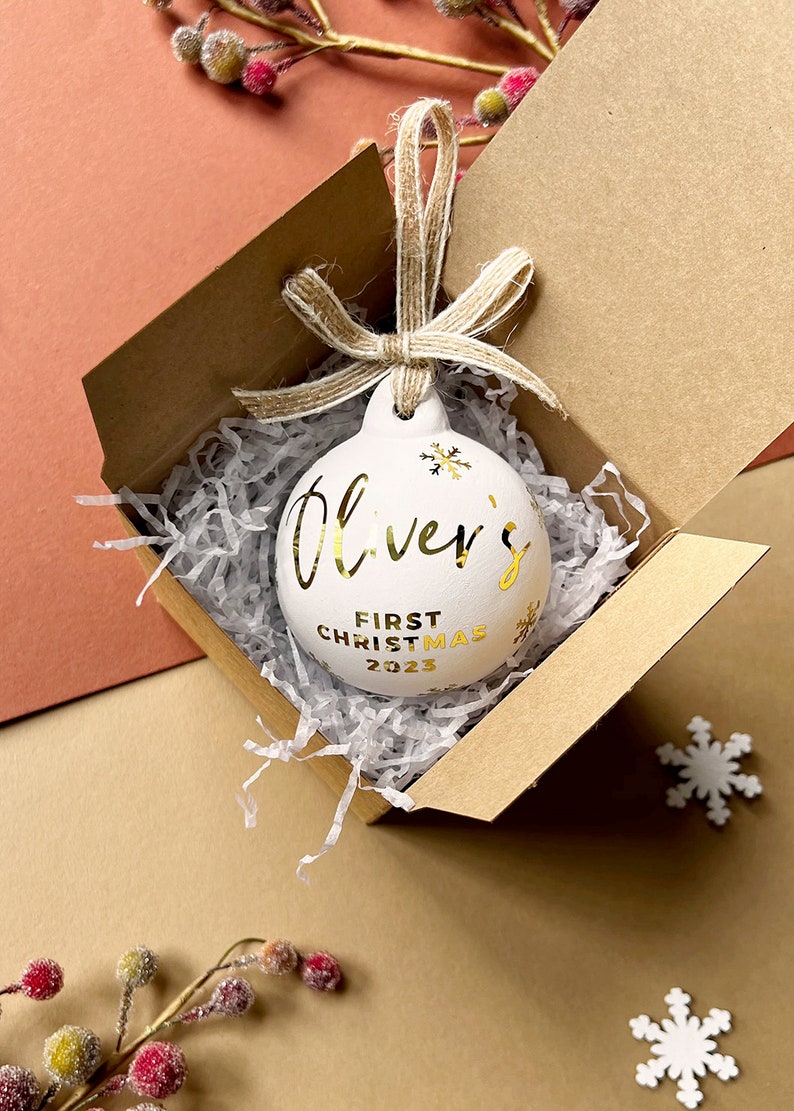May include: White Christmas ornament with gold lettering that reads "Oliver's First Christmas 2023." The ornament is in a brown box with shredded paper. A twine bow is attached to the ornament.