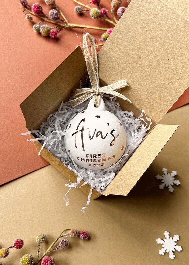 May include: A white Christmas ornament with the text "Ava's First Christmas 2023" in gold lettering. The ornament is inside a brown cardboard box with paper filler and a twine bow. The ornament has star cutouts.