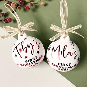 May include: Two white Christmas ornaments with red text and star accents. One ornament says "Toby's First Christmas 2024", the other says "Mila's First Christmas 2024". Each ornament has a twine bow.