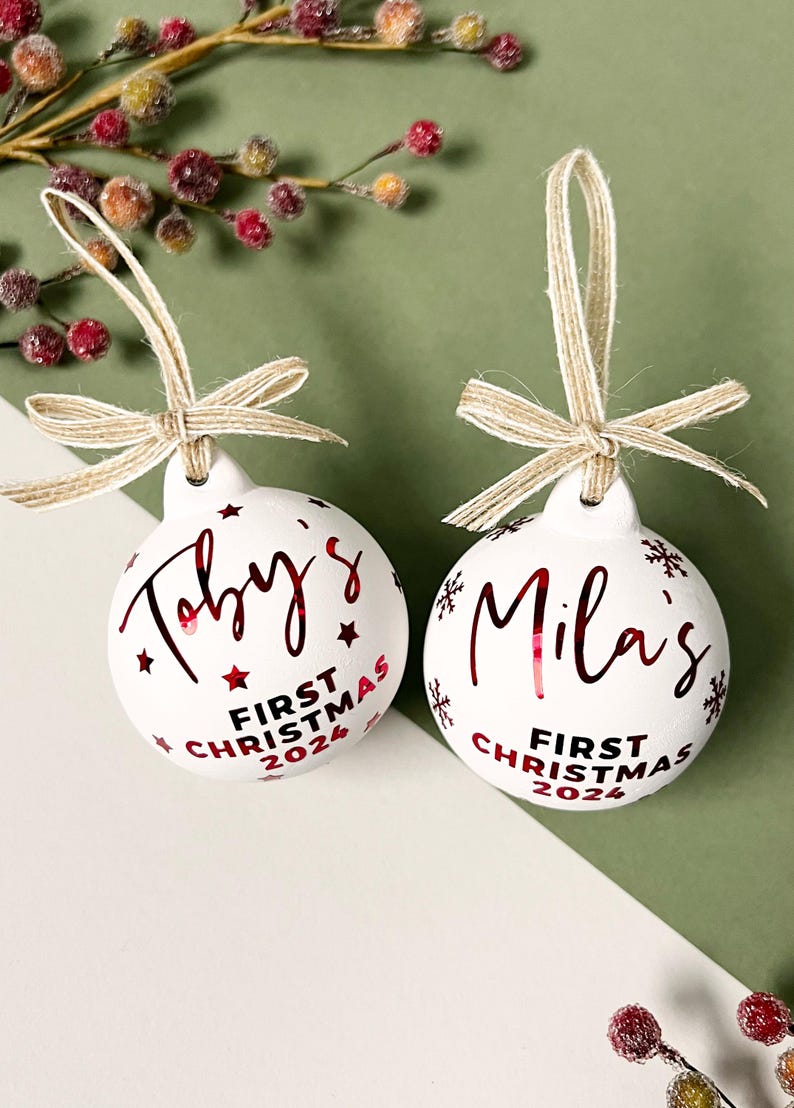 May include: Two white Christmas ornaments with red text and star accents. One ornament says "Toby's First Christmas 2024", the other says "Mila's First Christmas 2024". Each ornament has a twine bow.