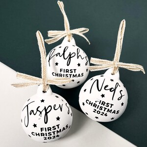 May include: Three white Christmas ornaments with black text and star accents. Each ornament has a name and "First Christmas 2024" printed on it. They are suspended by twine bows.