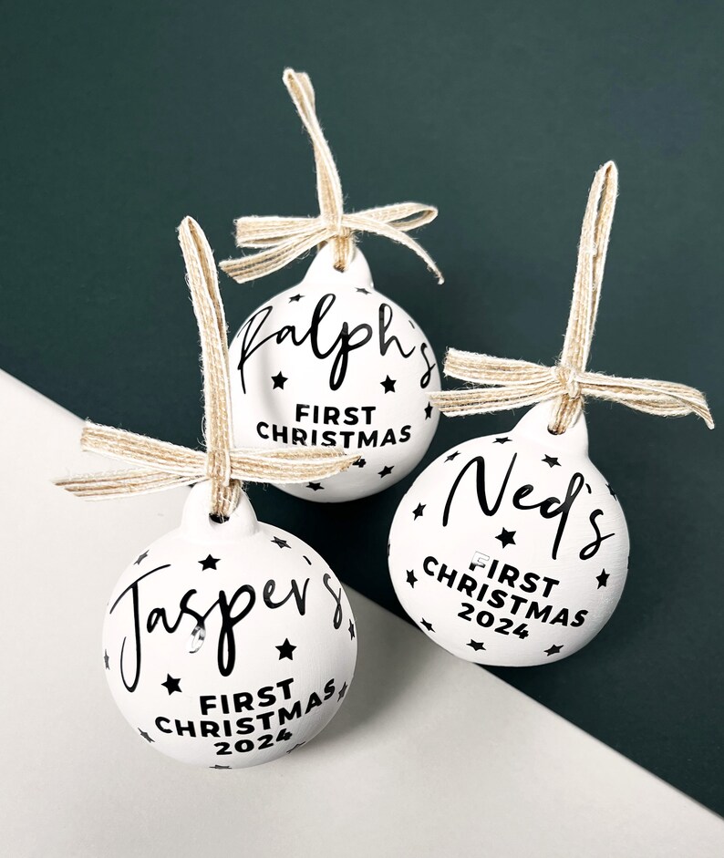 May include: Three white Christmas ornaments with black text and star accents. Each ornament has a name and "First Christmas 2024" printed on it. They are suspended by twine bows.