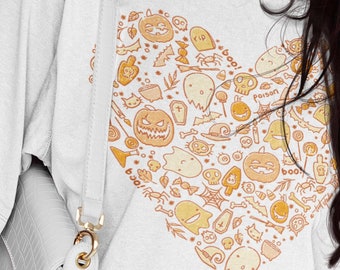 May include: White t-shirt featuring a heart-shaped design filled with Halloween-themed illustrations in orange and yellow. The design includes pumpkins, ghosts, and the word "boo!". A white handbag with a gold clasp is visible.