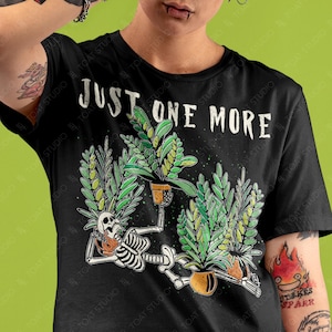 May include: Black t-shirt with the text "JUST ONE MORE" above a skeleton design holding potted plants. The graphic features green and yellow plants in orange pots. The shirt is a crew neck.