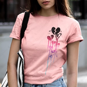 May include: Pink t-shirt featuring a graphic of broken hearts with a dripping effect in shades of pink, purple, and blue. The shirt has short sleeves and is paired with blue denim jeans. The text "TOAT STUDIO" is visible.