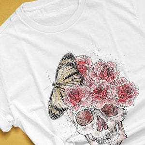 May include: White t-shirt featuring a skull design with a butterfly and roses. The skull is illustrated with red and gray shading, and the butterfly has black and yellow markings. The roses are pink. The text "colorway shown: WHITE" is visible.