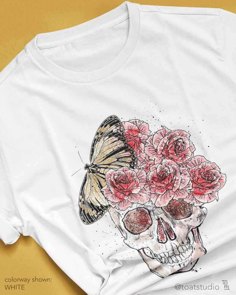 May include: White t-shirt featuring a skull design with a butterfly and roses. The skull is illustrated with red and gray shading, and the butterfly has black and yellow markings. The roses are pink. The text "colorway shown: WHITE" is visible.
