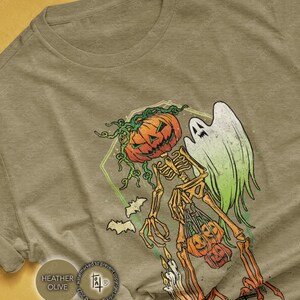 May include: Heather olive t-shirt featuring a Halloween-themed graphic. The design includes a skeleton with a pumpkin head, a ghost, bats, and pumpkins. The skeleton is holding pumpkins and candles. The shirt is a neutral color.