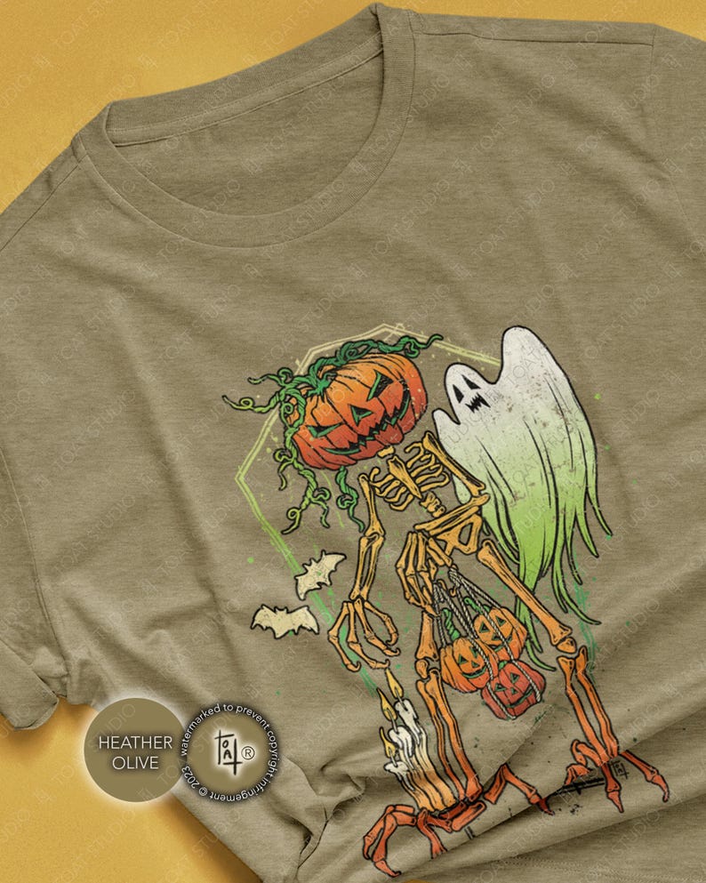 May include: Heather olive t-shirt featuring a Halloween-themed graphic. The design includes a skeleton with a pumpkin head, a ghost, bats, and pumpkins. The skeleton is holding pumpkins and candles. The shirt is a neutral color.