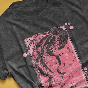 May include: A dark grey heather t-shirt with a pink and black graphic print of a jellyfish. The jellyfish is surrounded by pink splatter paint.