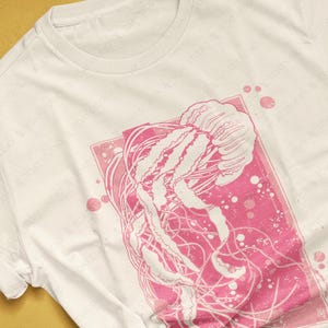 May include: A white t-shirt with a pink jellyfish graphic. The jellyfish is outlined in white and has a pink background. The graphic is surrounded by a pink square with white dots.