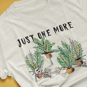 May include: A white t-shirt featuring the text "JUST ONE MORE" above a graphic of a skeleton tending to potted plants. The skeleton is surrounded by green plants in brown pots. The shirt is a natural color.
