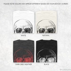 Pode incluir: Quatro camisetas dobradas em branco, natural, cinza escuro mesclado e preto. Cada camiseta apresenta um gr&aacute;fico detalhado de caveira. O texto "PLEASE NOTE COLORS MAY APPEAR DIFFERENT BASED ON YOUR DEVICE'S SCREEN" est&aacute; no topo.