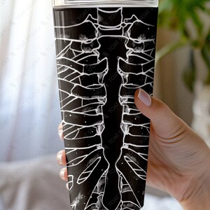 May include: A black and white stainless steel tumbler with a graphic of a human rib cage. The tumbler is held in a hand.