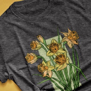 May include: A dark grey heather t-shirt with a graphic print of a bouquet of yellow daffodils with green leaves. The daffodils are drawn in a vintage style with a light green background.