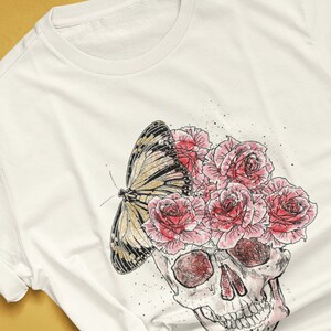 May include: Cream-colored t-shirt featuring a skull design with pink roses and a butterfly. The graphic is a watercolor style illustration. The text "colorway shown: NATURAL" is at the bottom left.