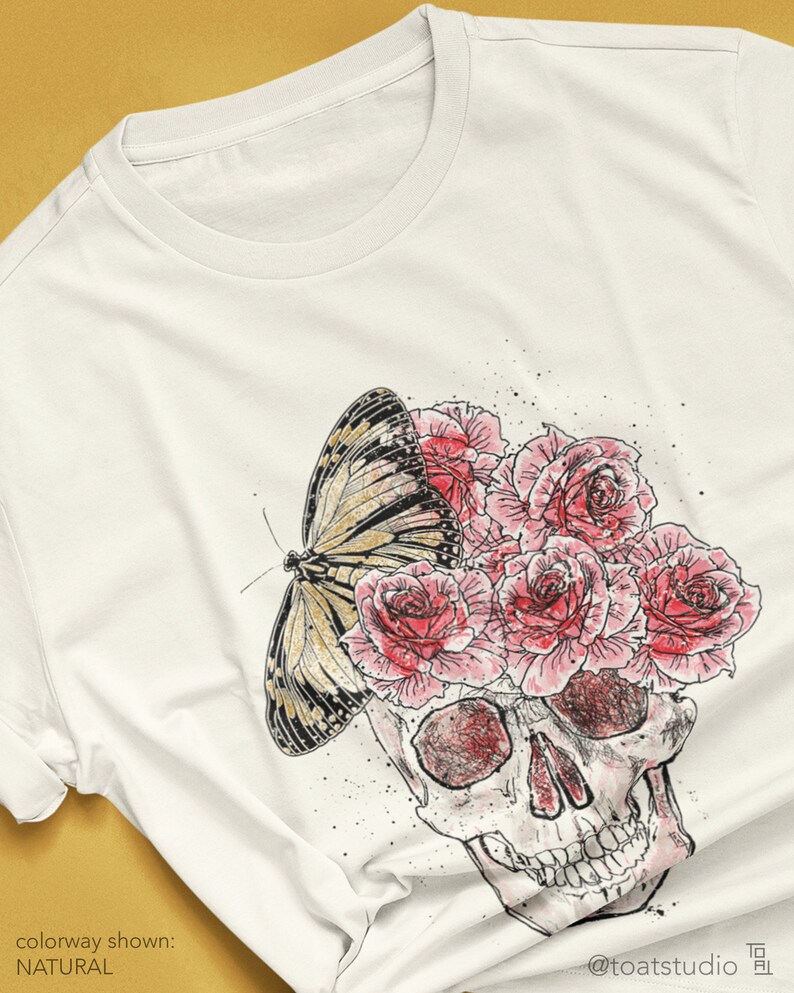 May include: Cream-colored t-shirt featuring a skull design with pink roses and a butterfly. The graphic is a watercolor style illustration. The text "colorway shown: NATURAL" is at the bottom left.
