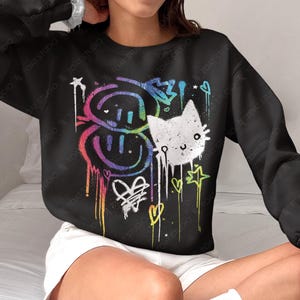 May include: Black sweatshirt with a colorful graphic design featuring smiley faces, a cat, hearts, and stars. The design has a graffiti-style aesthetic with dripping paint effects. The text "TOAT STUDIO" is visible.