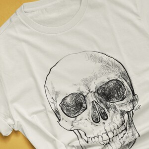 Pode incluir: Camiseta de cor natural com uma ilustra&ccedil;&atilde;o detalhada de caveira em preto e branco. O desenho da caveira &eacute; intrincadamente desenhado com linhas rabiscadas, criando um efeito texturizado. A combina&ccedil;&atilde;o de cores da camiseta &eacute; rotulada como "NATURAL". O nome do artista e seu handle s&atilde;o vis&iacute;veis.