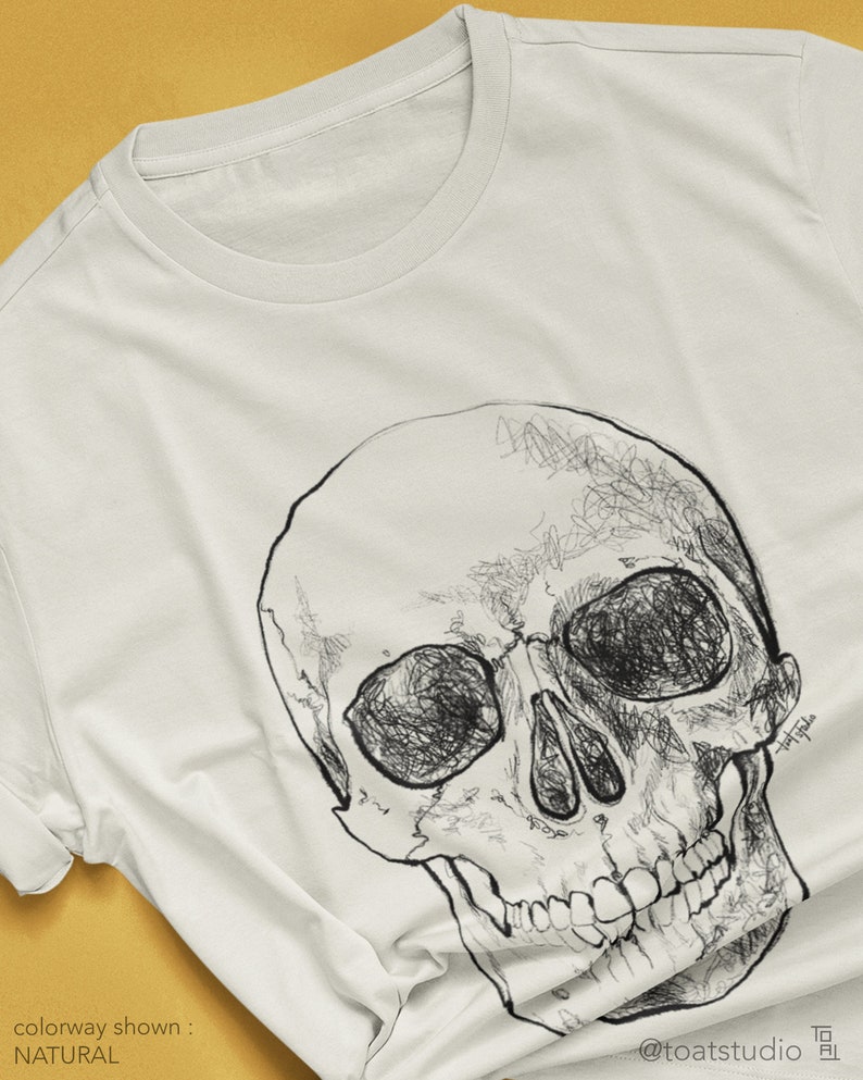 Pode incluir: Camiseta de cor natural com uma ilustra&ccedil;&atilde;o detalhada de caveira em preto e branco. O desenho da caveira &eacute; intrincadamente desenhado com linhas rabiscadas, criando um efeito texturizado. A combina&ccedil;&atilde;o de cores da camiseta &eacute; rotulada como "NATURAL". O nome do artista e seu handle s&atilde;o vis&iacute;veis.