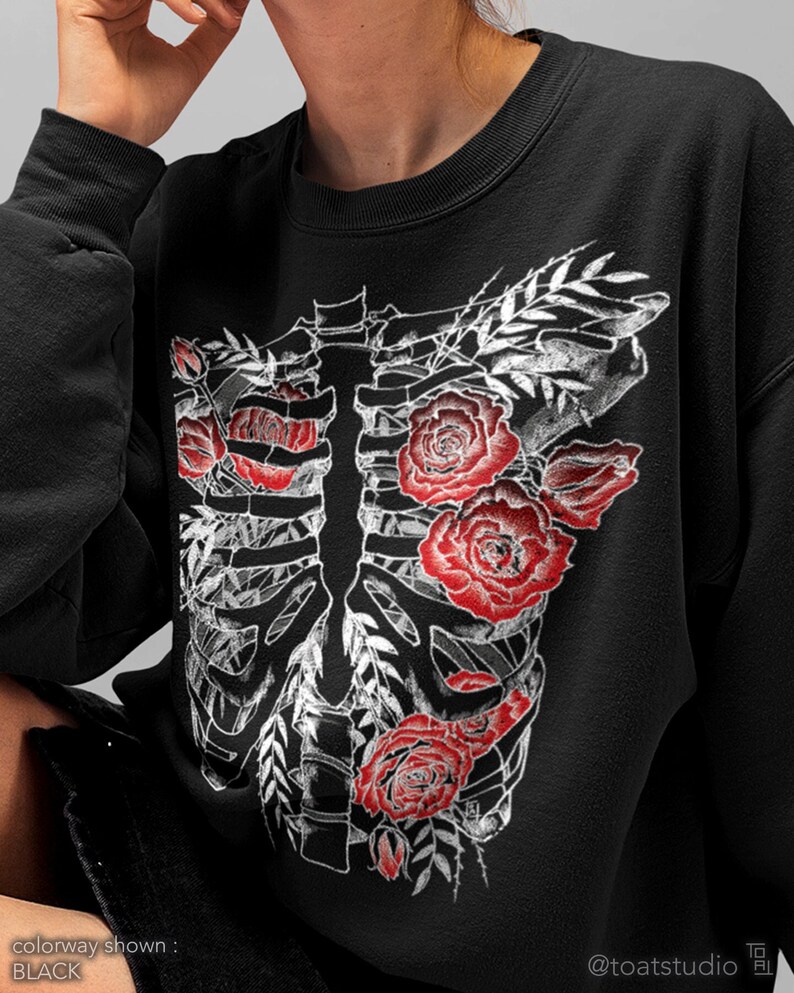 Pu&ograve; includere: Felpa nera con un design a gabbia toracica scheletrica bianca adornata da rose rosse e foglie bianche. Il testo "colorway shown: BLACK" &egrave; visibile in basso a sinistra. Il nome dell'artista, "@toatstudio", &egrave; in basso a destra.