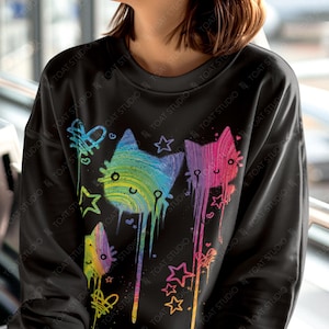 May include: Black sweatshirt with a colorful cat graphic. The design features three stylized cats in rainbow hues, dripping paint effects, and star accents. The text "colorway shown: BLACK" is at the bottom.