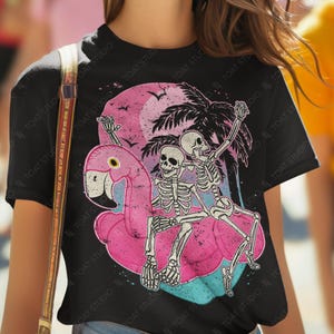 Op de afbeelding: Zwart T-shirt met een afbeelding van twee skeletten op een roze flamingo-drijver, tegen een roze zonsondergang en palmbomen. De tekst "TOAT STUDIO" is ook zichtbaar.