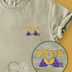 May include: Khaki-colored t-shirt with embroidered Japanese text and a sweet potato design. The shirt features a crew neck and short sleeves. The text and sweet potato design are in yellow and purple.