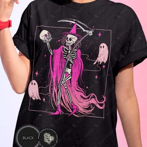 May include: Black t-shirt featuring a pink graphic of a skeleton in a witch hat holding a skull and scythe, with two pink ghosts. The design includes the text "TOAT STUDIO" and a copyright notice.
