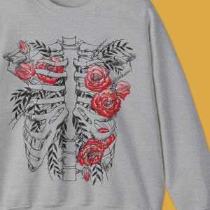 Pu&ograve; includere: Felpa grigio sport con un design a gabbia toracica floreale nera e rossa. La grafica include rose rosse e foglie nere. Il testo "colorway shown: SPORT GREY" &egrave; in basso nell'immagine, insieme al logo @toatstudio.