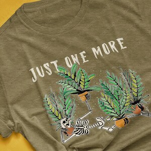 May include: Heather olive green t-shirt with the text "JUST ONE MORE" in white. The graphic features skeleton figures holding potted plants with green leaves. The shirt is a crew neck style.