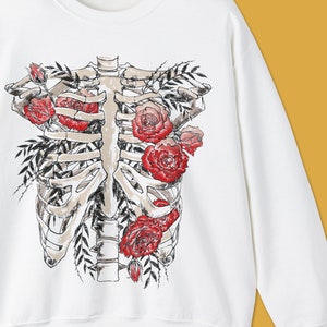 Pu&ograve; includere: Felpa bianca con grafica di gabbia toracica decorata con rose rosse e foglie verdi. Il disegno &egrave; centrato. Il testo "colorway shown : SPORT GREY" &egrave; nella parte inferiore dell'immagine.