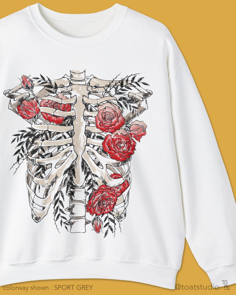 Pu&ograve; includere: Felpa bianca con grafica di gabbia toracica decorata con rose rosse e foglie verdi. Il disegno &egrave; centrato. Il testo "colorway shown : SPORT GREY" &egrave; nella parte inferiore dell'immagine.