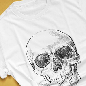 Pode incluir: Camiseta branca com uma ilustra&ccedil;&atilde;o de caveira em preto e branco. A caveira &eacute; desenhada com linhas detalhadas. O texto "colorway shown: WHITE" &eacute; vis&iacute;vel. O nome do artista e o handle tamb&eacute;m s&atilde;o vis&iacute;veis.