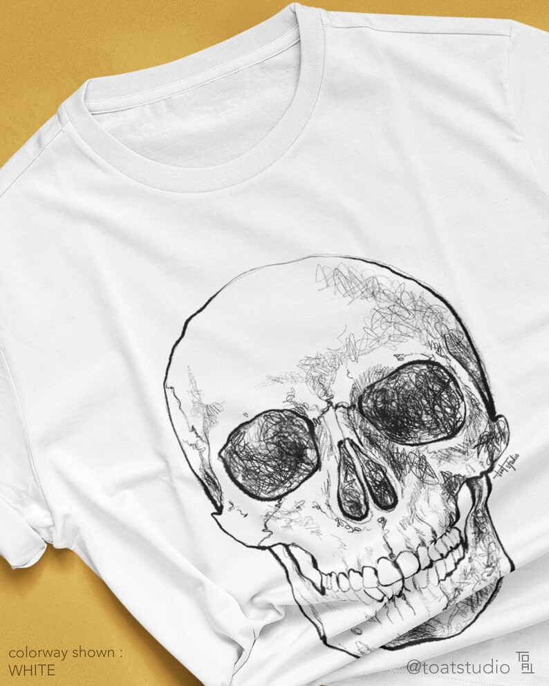 Pode incluir: Camiseta branca com uma ilustra&ccedil;&atilde;o de caveira em preto e branco. A caveira &eacute; desenhada com linhas detalhadas. O texto "colorway shown: WHITE" &eacute; vis&iacute;vel. O nome do artista e o handle tamb&eacute;m s&atilde;o vis&iacute;veis.