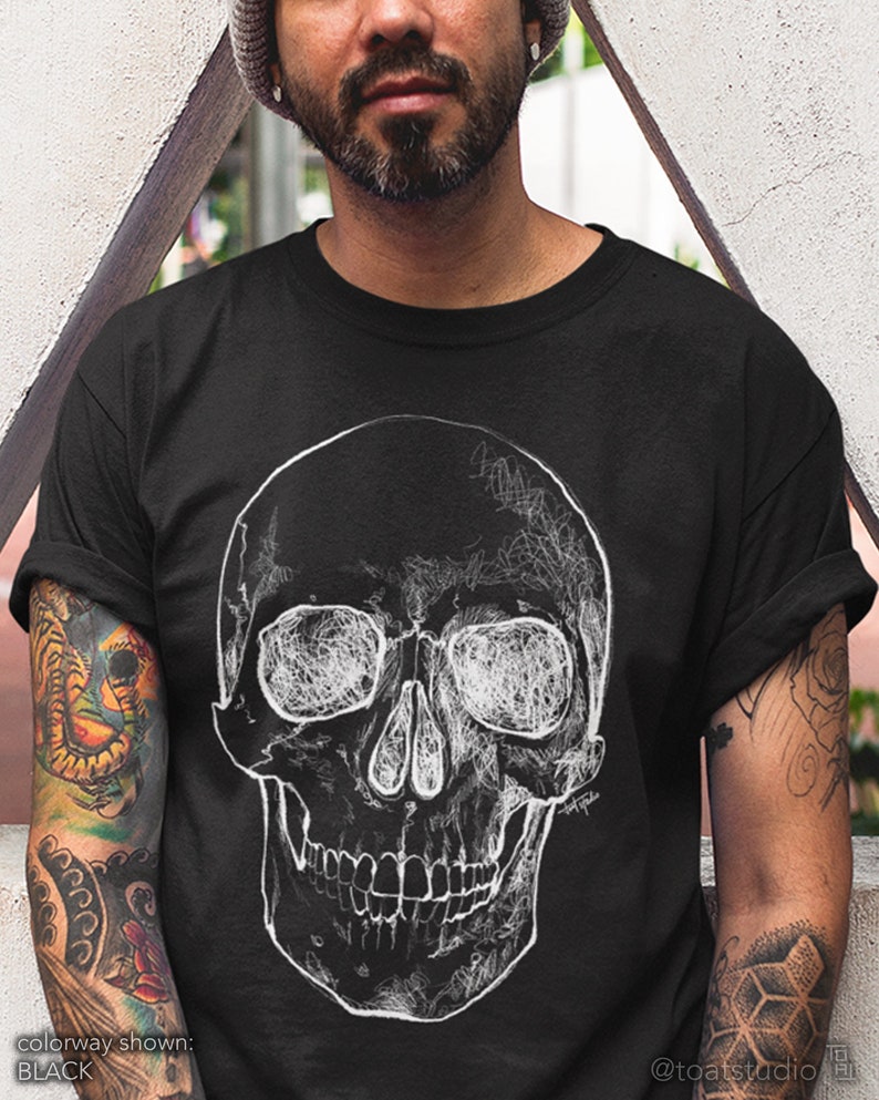 Pode incluir: Camiseta preta com um gr&aacute;fico de caveira branca detalhada. O design da caveira tem um estilo art&iacute;stico rabiscado. O texto "colorway shown: BLACK" est&aacute; na parte inferior esquerda. O logotipo @toatstudio est&aacute; na parte inferior direita.