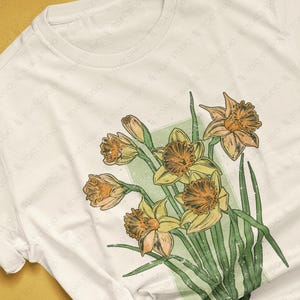 May include: A white t-shirt with a vintage-style illustration of a bouquet of yellow daffodils with green leaves. The design has a distressed, faded look.