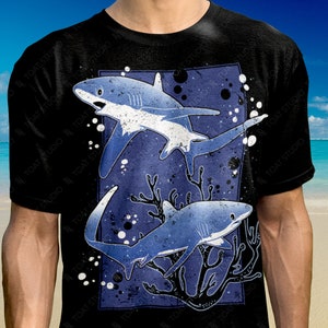 Op de afbeelding: Zwart T-shirt met een grafische print van twee blauwe haaien die zwemmen in een koraalrif. De haaien zijn gestileerd en hebben een witte onderbuik. Het koraalrif is donkerblauw met witte accenten. De achtergrond is donkerblauw met witte stippen.