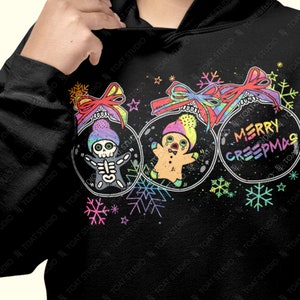 May include: Black hoodie featuring three Christmas ornament designs with spooky characters and the text "MERRY CREEPMAS". The ornaments include a skeleton, a gingerbread man, and colorful bows and snowflakes.