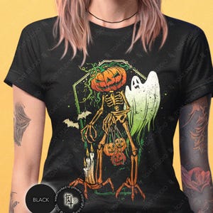 May include: Black Halloween-themed t-shirt featuring a skeleton with a pumpkin head, holding pumpkins and candles. A green ghost and bats are also in the design. The shirt has the text "TOAT STUDIO" printed on it.