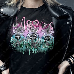 May include: Black t-shirt featuring a graphic of three cartoon plush toys: a bunny, a bear, and a cat, each with a skeleton design. The design includes pink, blue, and purple accents. The text "colorway shown: BLACK" is at the bottom.
