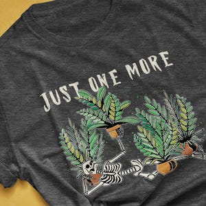 May include: Dark grey heather t-shirt with the text "JUST ONE MORE" above a graphic of skeletons holding potted plants. The skeletons are surrounded by green plants and brown pots. The shirt's colorway is dark grey heather.