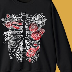 Pu&ograve; includere: Felpa nera con un design a gabbia toracica scheletrica bianca adornata da rose rosse e foglie verdi. Il testo "colorway shown: BLACK" &egrave; in basso a sinistra.
