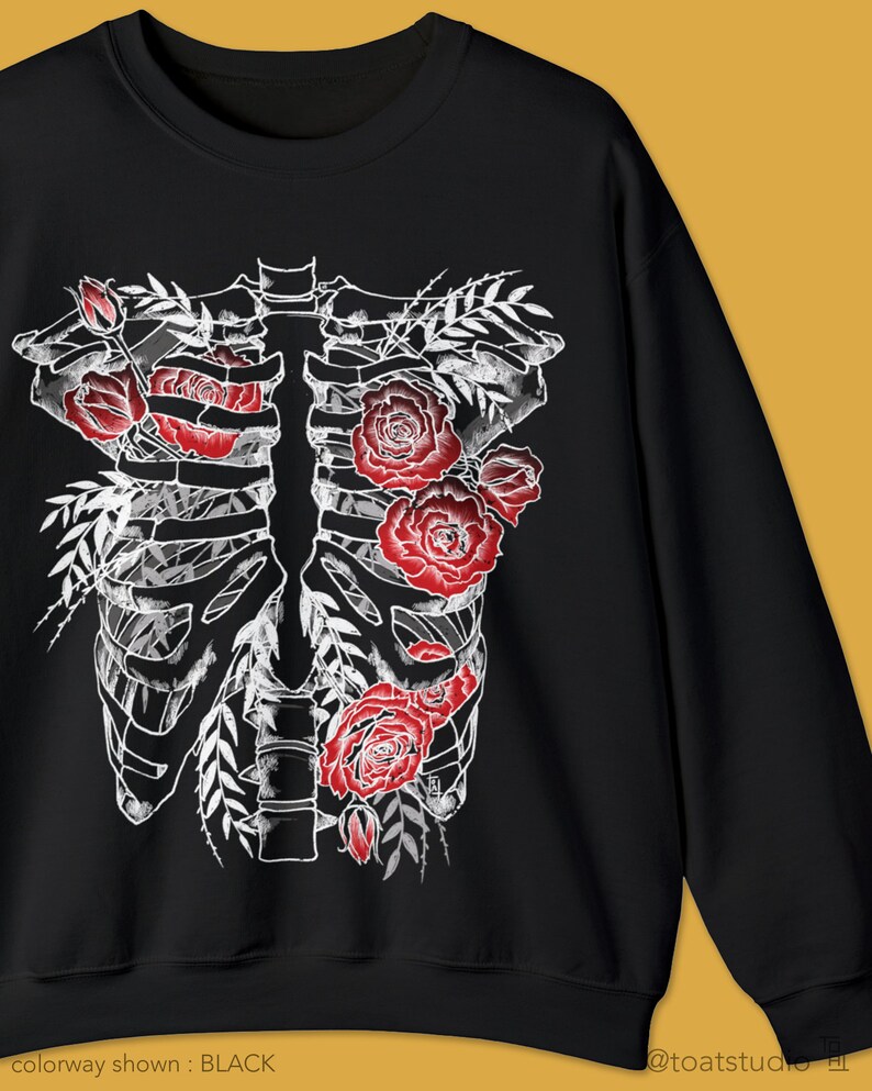 Pu&ograve; includere: Felpa nera con un design a gabbia toracica scheletrica bianca adornata da rose rosse e foglie verdi. Il testo "colorway shown: BLACK" &egrave; in basso a sinistra.