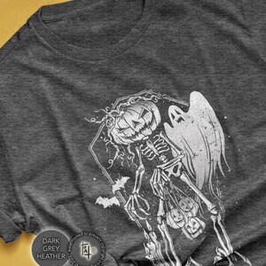 May include: Dark grey heather t-shirt featuring a white Halloween design. The graphic includes a skeleton with a pumpkin head, a ghost, and bats. The text "DARK GREY HEATHER" is printed on the shirt.