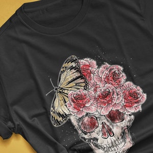 May include: Black t-shirt featuring a graphic design of a skull with pink roses and a butterfly. The butterfly has yellow and black markings. The text "colorway shown: BLACK" is visible. The artist's name is also visible.