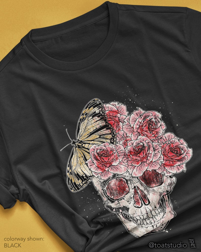 May include: Black t-shirt featuring a graphic design of a skull with pink roses and a butterfly. The butterfly has yellow and black markings. The text "colorway shown: BLACK" is visible. The artist's name is also visible.
