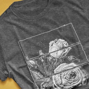 May include: Dark grey heather t-shirt featuring a detailed black and white rose graphic. The design includes a stylized illustration of roses and buds within a rectangular frame. The text "colorway shown: DARK GREY HEATHER" is visible.