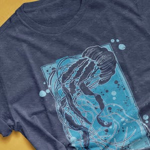 May include: A heather navy blue t-shirt with a graphic print of a jellyfish in blue and white. The jellyfish is surrounded by blue and white dots.