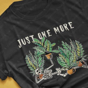 May include: Black t-shirt with a graphic featuring a skeleton reclining with potted plants, and the text "JUST ONE MORE". The design includes green foliage and brown pots. The shirt's colorway is black.